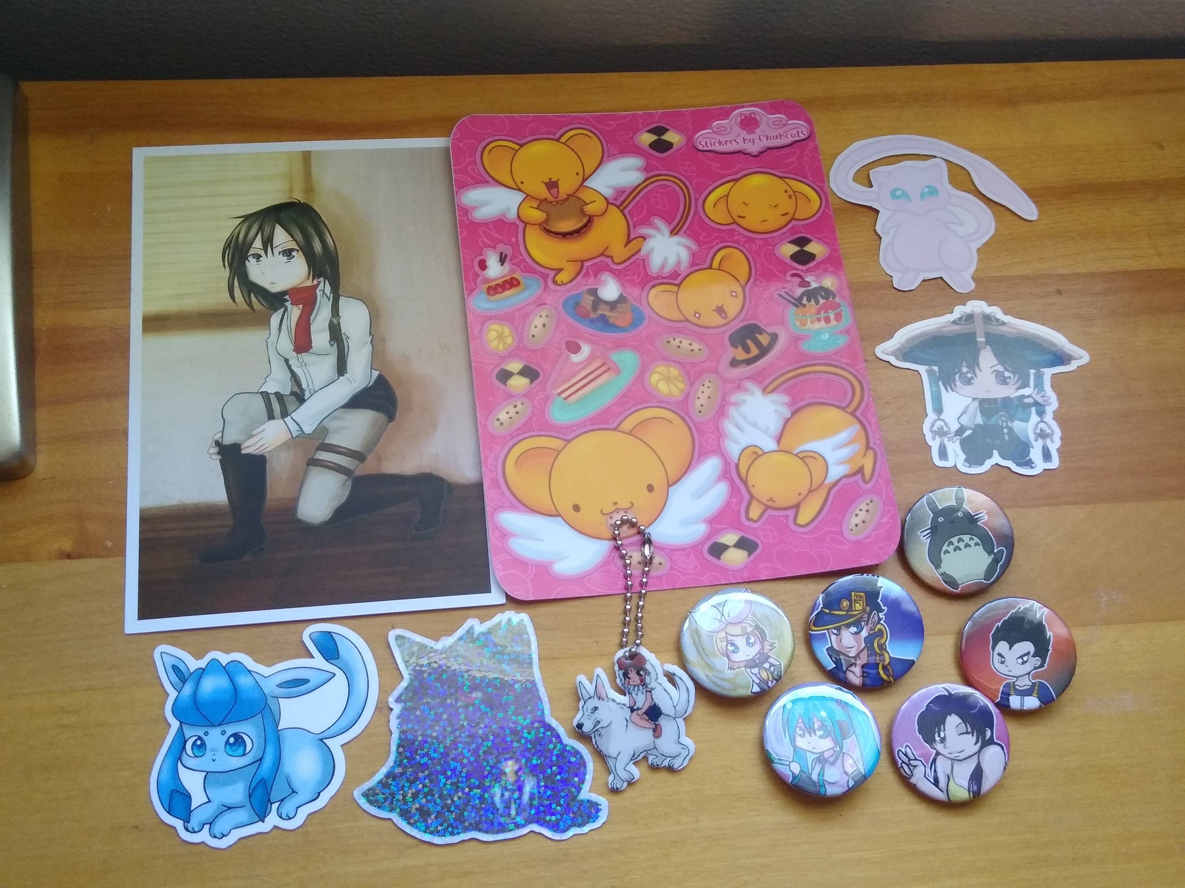 A selection of merch from Chubcast, laid out atop a maple-wood desk. From left to right/top to bottom, there is: A print of Mikasa from Attack on Titan kneeling and putting on a boot, a sticker sheet of Kero from Cardcaptor Sakura amidst a variety of sweets, a sticker of Mew from Pokemon, a sticker of the Wanderer from Genshin Impact, a sticker of a Vaporeon, a sticker of a Gengar with a Beedrill in the jar in its hands, a charm of San and Moro from Princess Mononoke, and button pins of: Kagamine Rin and Hatsune Miku from Vocaloid, Jotaro Kujo from JoJo's Bizarre Adventure, Totoro from My Neighbor Totoro, Misato Katsuragi from Neon Genesis Evangelion, and Vegeta from Dragon Ball Z.