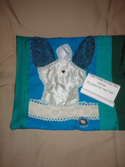 panel 1 applique angel silver-blue fabric and black wings, on lace seafoam with applique fish and scroll with words Don't step on that fish Castiel. Big plans for that fish.