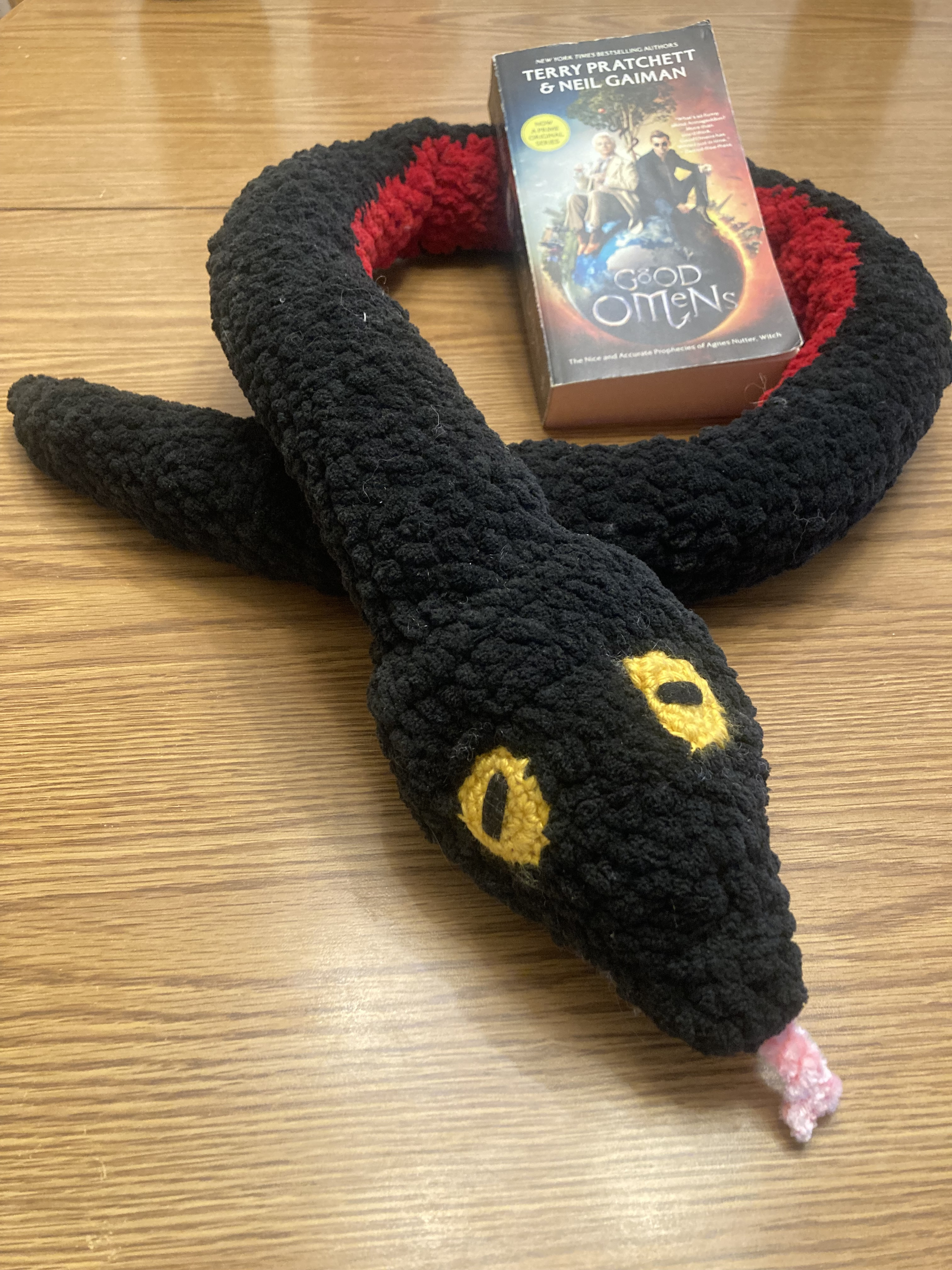 A black amigurumi snake wrapped around a copy of the novel Good Omens.