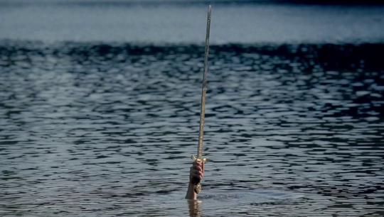 a shot of a hand rising from the lake holding a sword