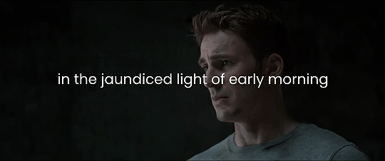 a gif of Steve staring at Bucky in the warehouse after he talked about Steve wearing newspaper in his shoes and the text on the gif reads in the jaundiced light of early morning.