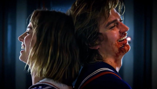 a side shot of Robin and Steve tied up with their backs to each other smiling as they laugh even though Steve has blood on his face