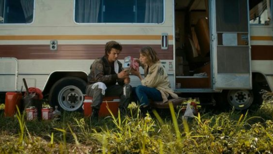 zoomed out shot of Steve and Robin in the field as Steve holds a bottle and Robin pours gasoline into the bottle.