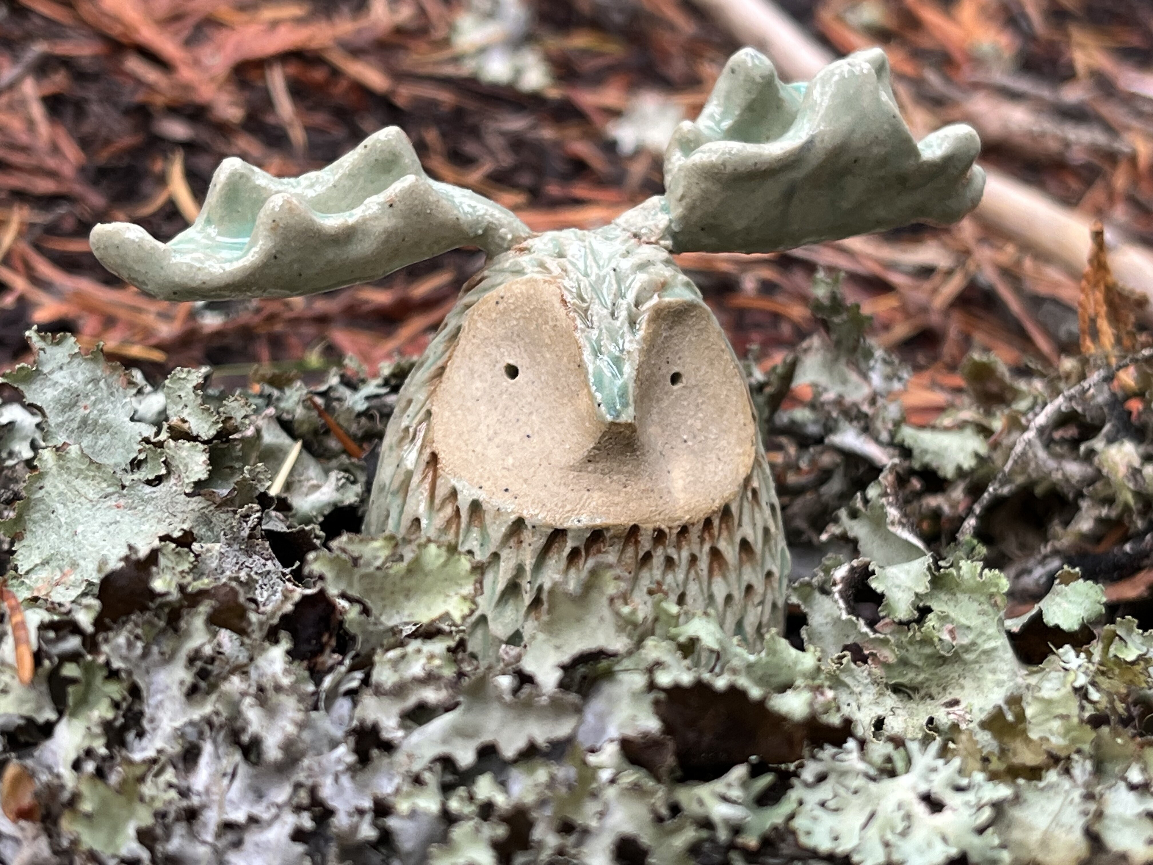An image of a small ceramic creature with antlers in a forest, nested in pale lichen.